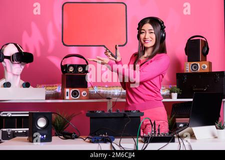 Lächelnder Künstler, der im Studio eine Sprechblase in roter Schachtel mit isolierten Textnachrichten aus Pappe wirbt. Junge dj-Frau mit Plakatvorlage, Kosmetikraum, Plattenspieler aus Vinyl. Stockfoto