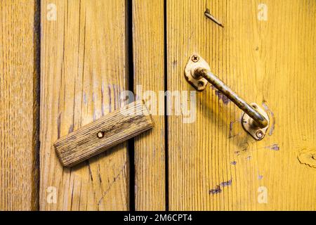 Die zerkratzte Tür des alten Dorfes mit einem rostigen, gelb lackierten Griff ist auf einem Holzschloss verschlossen. Stockfoto