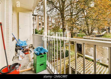 Ein Balkon mit Mülleimern und Mopps auf dem Boden neben einer offenen Tür, vor einem Baum Stockfoto