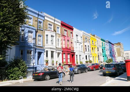 Ein junges Paar, das an einem sonnigen Tag Händchen hält und eine Straße mit bunten Reihen von Reihenhäusern überquert, Notting Hill, London, England, Großbritannien