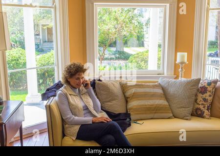 NEW ORLEANS, LA, USA - 5. NOVEMBER 2021: Seniorin sitzt auf dem Sofa im Wohnzimmer, während sie ihr Smartphone benutzt Stockfoto
