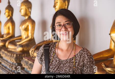 Porträt einer asiatischen Touristin in einem thailändischen Tempel, Frau mittleren Alters. Trägt eine Brille, lächelt und schaut in die Kamera, verschwommener Hintergrund oder Stockfoto