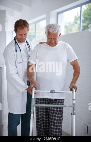 Ich komme langsam dorthin. Ein männlicher Arzt, der seinem älteren Patienten assistiert, der eine Gehhilfe benutzt. Stockfoto