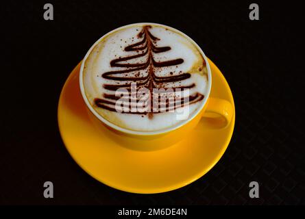 Schaumiger Cappuccino. Isolierter gelber Becher. Dekorative Weihnachtsbaumschmuck auf weißem Schaum. Gourmet-Kaffee. Brauner Schokoladenkiefernbaum. Schwarzer Hintergrund Stockfoto