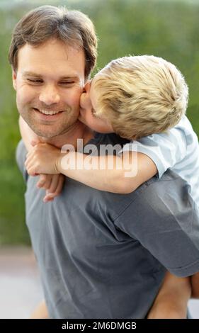 Die Liebe zu einem Sohn. Der Junge küsst seinem Vater die Wange, während er ihm Huckepack gibt. Stockfoto