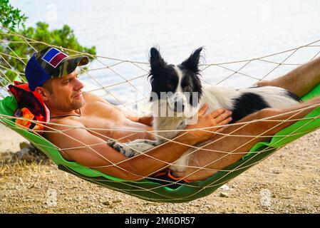 Ein junger Mann ruht sich mit einem Hund in einer Hängematte am Strand aus. Stockfoto