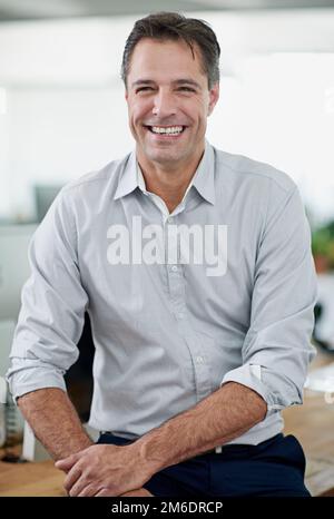 Er ist ein umsichtiger Manager. Porträt eines erwachsenen Geschäftsmanns, der auf seinem Schreibtisch sitzt. Stockfoto