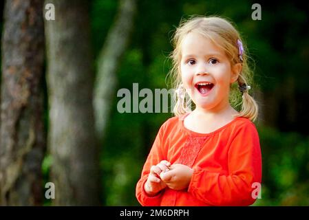 Ein kleines, 3 Jahre altes, fröhliches Mädchen sitzt auf einem Holzstamm im Wald. Stockfoto