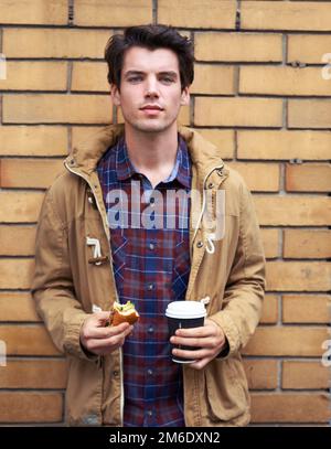 Nahrung für Geist, Körper und Seele. Ein junger Mann steht draußen mit einem halb gefressenen Burger und Kaffee zum Mitnehmen. Stockfoto