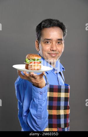 Indischer männlicher Koch mit einem Burger in der Hand auf grauem Hintergrund Stockfoto