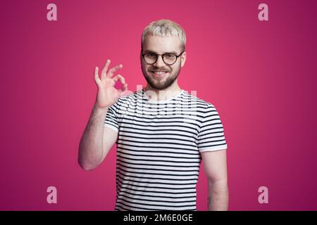 Glücklicher junger Mann, der ein T-Shirt trägt, das OK-Zeichen zeigt und vor pinkfarbenem Hintergrund steht. Stockfoto