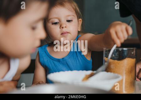 Das niedliche Mädchen, das Mehl in die Schüssel mit Teigzutaten gibt, hilft der Mutter dabei, leckere Muffins in der Küche zuzubereiten. Backen mit Kindern Stockfoto