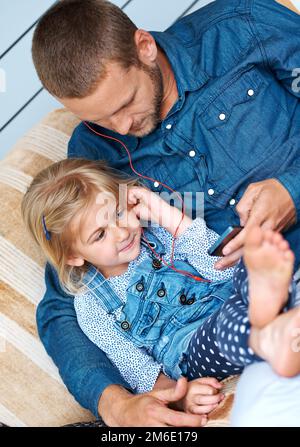 Sie wählen ihren nächsten Titel. Ein bezauberndes kleines Mädchen, das auf dem Sofa sitzt und mit seinem Vater Kopfhörer teilt. Stockfoto