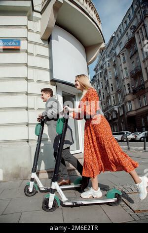 Junge liebende Paar entspannen sitzen in der Nähe eines modernen Glasgebäude mit ihren Elektro-Scooter. Spaziergänge in der großen Stadt. Stockfoto