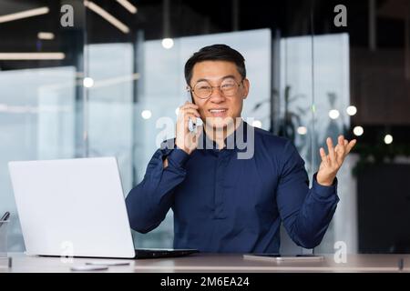 Erfolgreicher und fröhlicher asiatischer Geschäftsmann, der mit Kunden im Büro telefoniert, ein erwachsener Mann in einem Hemd, der am Arbeitsplatz an einem Laptop arbeitet. Stockfoto
