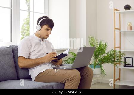 Junger Mann, der auf dem Sofa sitzt, online lernt, sich einen Laptop ansieht und sich Notizen macht Stockfoto