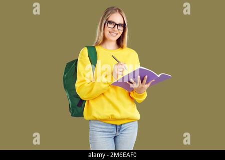 Lächelndes blondes Mädchen mit Brille und Rucksack macht Notizen im Copybook auf Khaki-Hintergrund. Stockfoto