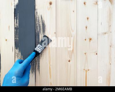 Die Hand eines Arbeiters in blauen Gummihandschuhen bemalt eine Holzwand mit einem Pinsel grauer Farbe. Reparieren Sie die Oberfläche. Stockfoto