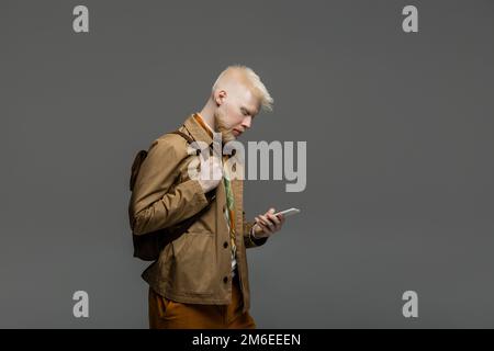 Bärtiger Albino-Mann, der mit Rucksack steht und ein Smartphone benutzt, isoliert auf grauem Stockbild Stockfoto