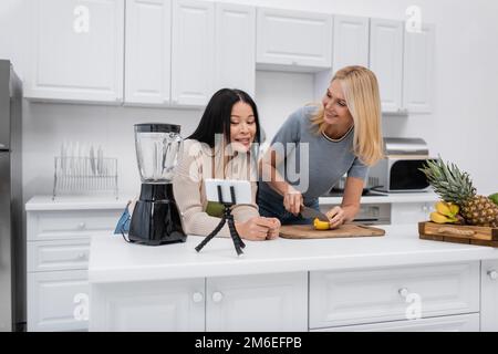 Lächelnde asiatische Frau, die sich in der Nähe des Smartphones auf dem Stativ unterhält, während ein Freund in der Küche Obst schneidet, Stockbild Stockfoto