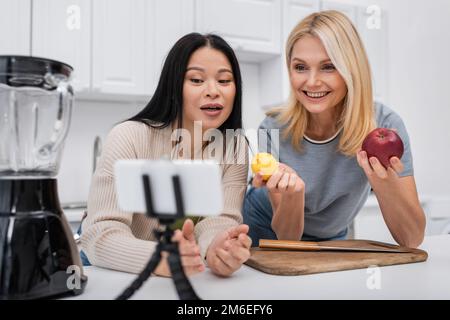 Fröhliche Frau mit Früchten, während ein asiatischer Freund mit einem Smartphone auf einem Stativ in der Küche spricht, Stockbild Stockfoto