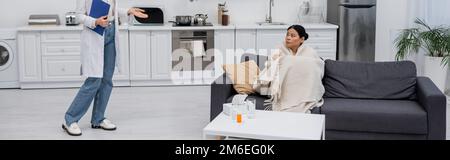 Eine kranke asiatische Frau, die zu Hause einen Arzt ansieht, mit einem Klemmbrett, einem Banner, einem Stockbild Stockfoto