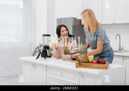 Lächelnde asiatische Frau, die Zitrone hält und mit einem Freund spricht, der Apfel in der Nähe des Smartphones schneidet, auf einem Stativ in der Küche, Stockbild Stockfoto