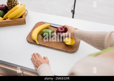 Zugeschnittene Ansicht einer Frau, die einen Apfel neben dem Schneidebrett auf dem Tisch hält, Stockbild Stockfoto
