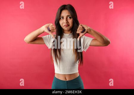 Eine junge weiße Frau trägt ein lässiges Oberteil isoliert auf rotem Hintergrund und macht mit beiden Händen eine Daumen-nach-unten-Geste. Nicht zufrieden und schaut auf das Kommen Stockfoto