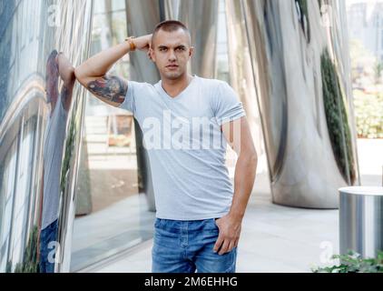 Ein junger Mann lehnt sich auf eine glänzende Säule. Mutiger Kerl mit einer Feuerwehrtätowierung auf seinem Bizeps. Männliches Porträt. Stockfoto