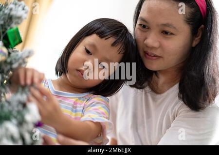 Ein kleines asiatisches Mädchen mit mom, die zusammen einen Weihnachtsbaum schmückt Stockfoto