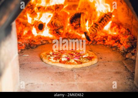 Pizza mit Belag in einem heißen Steinofen mit Feuerholz, das eine köstliche Mahlzeit backt Stockfoto