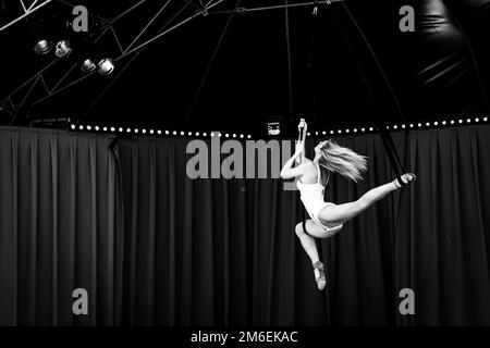 Ein elegantes Schwarzweiß-Nahporträt eines Zirkus-Entertainers, der hoch oben in einer wunderschönen Pose hängt und Seile in einem Zelt benutzt. Die flexible artis Stockfoto