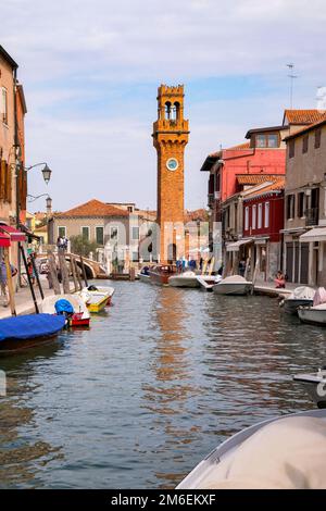 Fischerboote und bunte venezianische Häuser entlang des Kanals auf den Inseln Murano, mit Torre dell'Orologio im Hintergrund Stockfoto