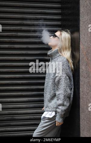 Seitenansicht einer jungen Frau in Winterpullover und Sonnenbrille, die E-Zigarette raucht, während sie auf der Straße der Stadt steht, Stockbild Stockfoto