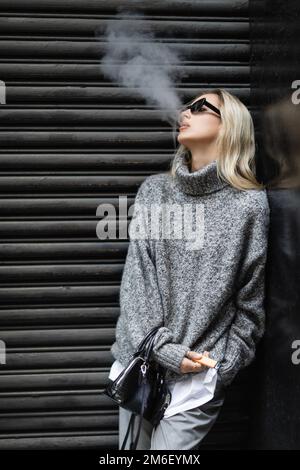 Junge Frau in Winterpullover und Sonnenbrille, die E-Zigarette raucht, während sie mit Handtasche auf der Straße steht, Stockbild Stockfoto