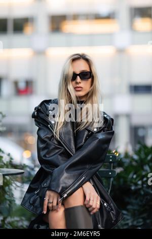 Junge blonde Frau in schwarzer Lederjacke und Sonnenbrille, sitzt im Café im Freien, Stock Image Stockfoto