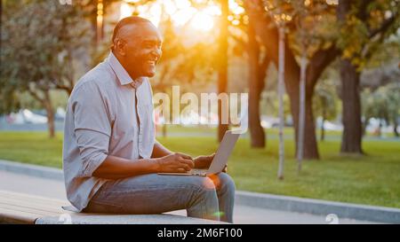 Ein fröhlicher, sorgenfreier, hochrangiger afroamerikanischer Mann, der im Freien sitzt, arbeitet mit einem Laptop und spielt Online-Spiele, hat Spaß beim Tippen auf dem Computer. Ethnisch reif Stockfoto