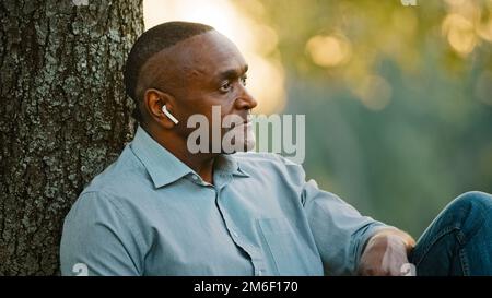 Porträt mit Kopfschuss Nahaufnahme ältere Erwachsene im mittleren Alter Afroamerikaner Geschäftsmann entspannt unter einem Baum im Stadtpark sitzen Arbeitspause zuhören Stockfoto