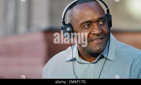 Nahaufnahme eines Mannes mittleren Alters, emotionaler, hochrangiger afroamerikanischer Mann, der sich im Freien ausruht und ein Lied über Kopfhörer hört und dabei großartigen Sound genießt Stockfoto