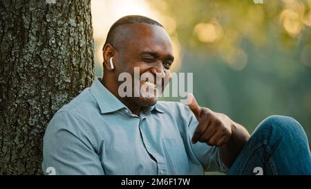 Porträt älterer afroamerikanischer älterer erwachsener Mann mittleren Alters, der im Park unter einem Baum sitzt und sich im Freien ausruht und Musik hört Stockfoto