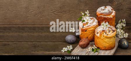 Osterkuchen Cruffin oder süßes Brot kulich mit bemalten Eiern. Traditionelles festliches Backen und frische Kirschblumen, festlicher Holzhintergrund. Jahrgang b Stockfoto