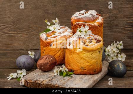 Osterkuchen Cruffin oder süßes Brot kulich mit bemalten Eiern. Traditionelles festliches Backen und frische Kirschblumen, festlicher Holzhintergrund. Jahrgang b Stockfoto