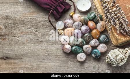 „Elder Futhark Rune Stones“-Set aus natürlichen Edelsteinen mit altem Buch, trockenen Kräutern und Salbei Incense auf Holzhintergrund mit Kopierbereich Stockfoto