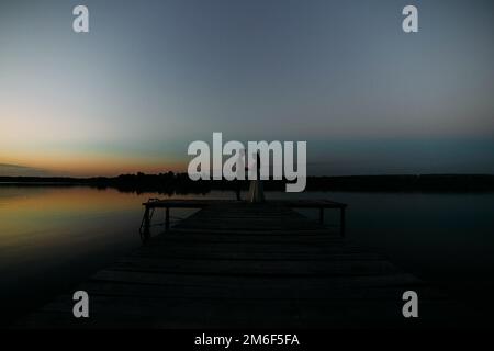 Braut und Bräutigam stehen bei Sonnenuntergang auf einem hölzernen Pier. Stockfoto