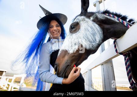 Ein Mädchen als Hexe gekleidet steht neben einem Pferd, auf das ein Skelett für die Feier gezogen wird Von Halloween Stockfoto