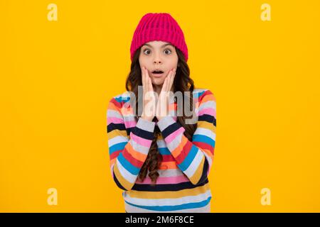 Wunderschönes Winterporträt für Kinder. Teenager-Mädchen mit Winterpullover und Strickmütze auf gelbem Hintergrund. Schockiertes Gesicht, überraschende Emotionen Stockfoto