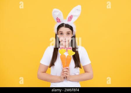 Hasenmädchen mit Spielzeugmöhren. Hübsches Mädchen mit Hasenohren isoliert auf gelbem Hintergrund. Die Osterhasen-Frau sieht lustig aus. Stockfoto