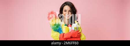 Fröhliche braune Hausfrau mit farbenfrohem Staubwedel und Reinigungsmittel, die auf die Kamera schauen, isoliert auf pinkfarbenem Banner, Stock-Bild Stockfoto