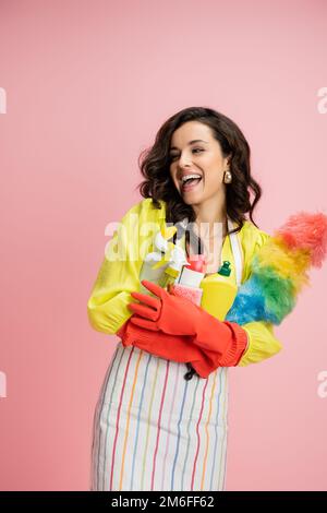 Lachende braune Frau in roten Gummihandschuhen mit farbenfrohem Staubwedel und Reinigungsmittel isoliert auf pinkfarbenem Stockbild Stockfoto